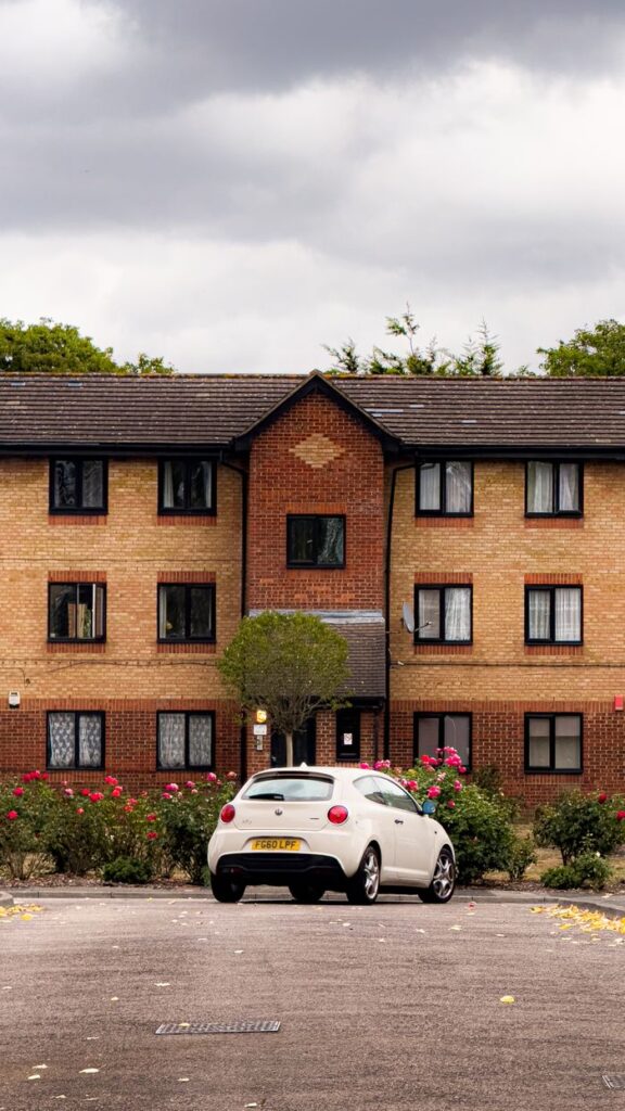 Brick apartment building with a white car parked, cloudy sky, and rose bushes.